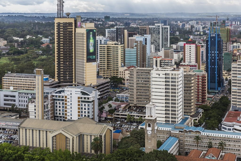 Aerial view of a dense urban skyline with a mixture of modern high-rise buildings and older architectures, prominent clock tower in the foreground, and a backdrop of distant trees and overcast skies.