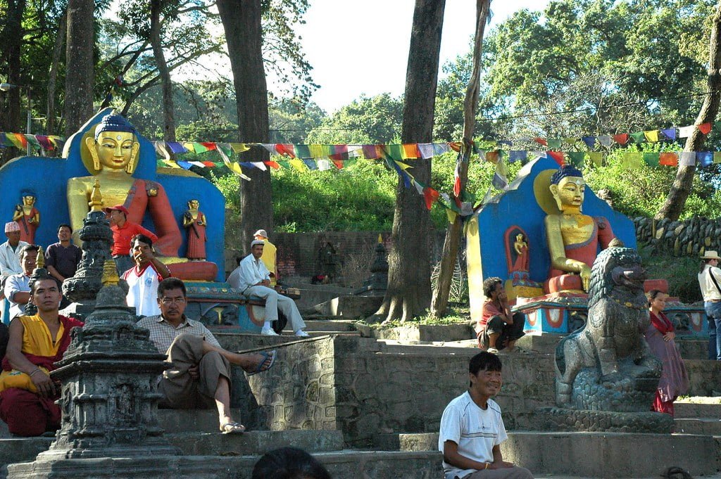 Alt text: People relax and engage in conversation in front of two large, colorful Buddha statues adorned with golden paint. Prayer flags flutter above in a serene outdoor setting with trees.
