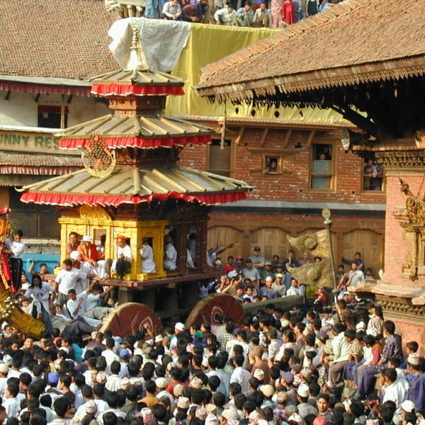 A vibrant cultural festival with a large crowd gathering around a traditional Nepalese chariot procession in a historic square, with intricately carved wooden architecture and onlookers observing from balconies.