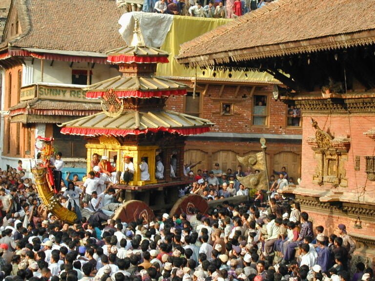 A vibrant cultural festival with a large crowd gathering around a traditional Nepalese chariot procession in a historic square, with intricately carved wooden architecture and onlookers observing from balconies.