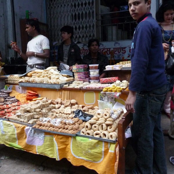 A busy street food stall with various types of sweets and snacks displayed on a table, attended by a vendor talking to a customer, with other people browsing and buying food.