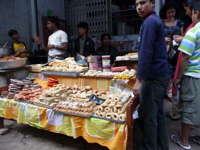 A busy street food stall with various types of sweets and snacks displayed on a table, attended by a vendor talking to a customer, with other people browsing and buying food.