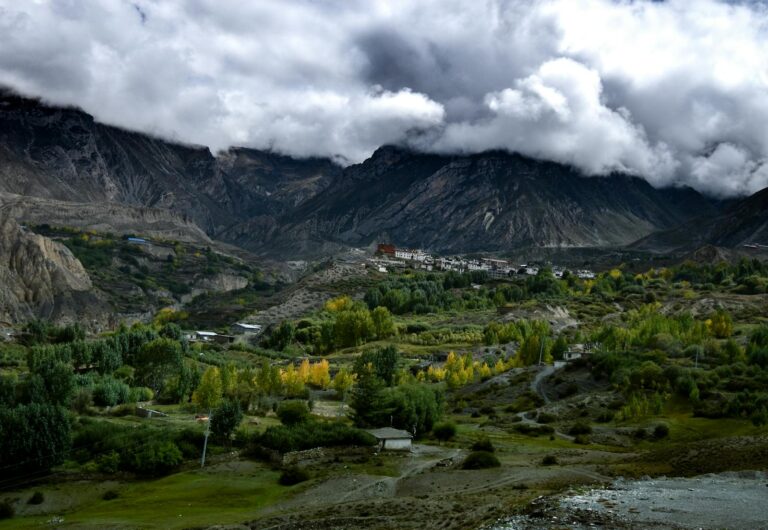 A small village nestled in a mountainous landscape with autumnal trees in the valley under a cloudy sky.