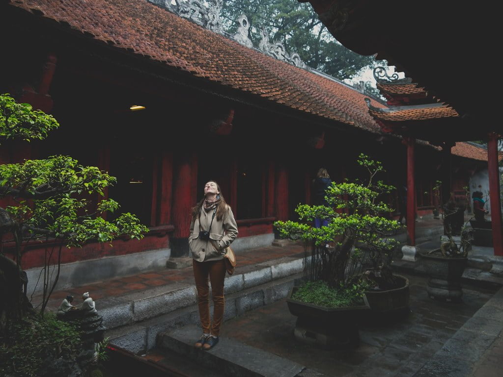 A person standing in an ancient temple courtyard with bonsai trees and traditional red columns and tiled roofs, looking upwards with a contemplative expression.