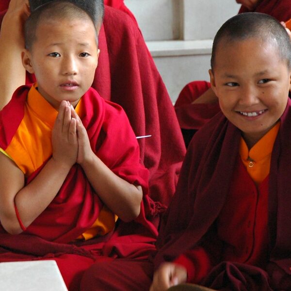 Two young monks in traditional red and yellow robes smiling and posing with one making the Anjali Mudra (prayer hands).