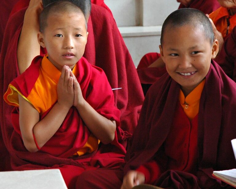 Two young monks in traditional red and yellow robes smiling and posing with one making the Anjali Mudra (prayer hands).