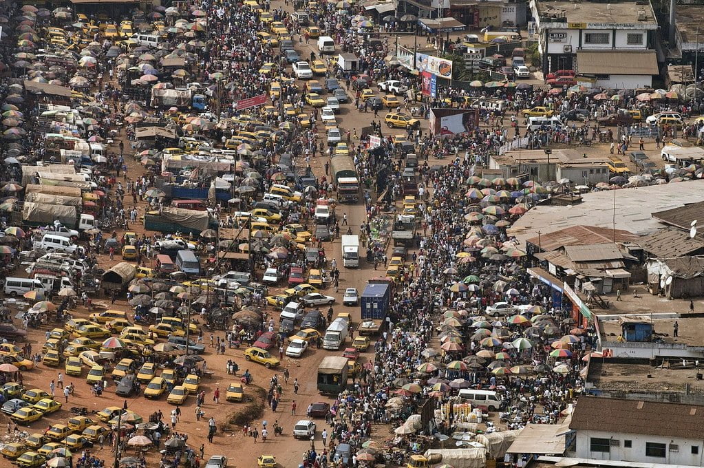 A busy street scene in a densely populated urban area with numerous people and colorful umbrellas, surrounded by a mix of vehicles and bustling market activity.