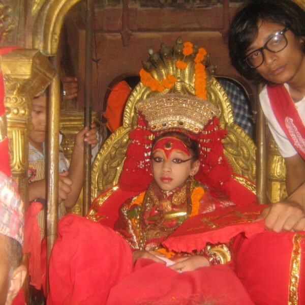 Alt text: A young girl dressed in elaborate traditional attire with a red and gold headdress sits on a golden palanquin, surrounded by attendants, during a cultural ceremony.