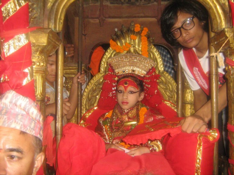 Alt text: A young girl dressed in elaborate traditional attire with a red and gold headdress sits on a golden palanquin, surrounded by attendants, during a cultural ceremony.