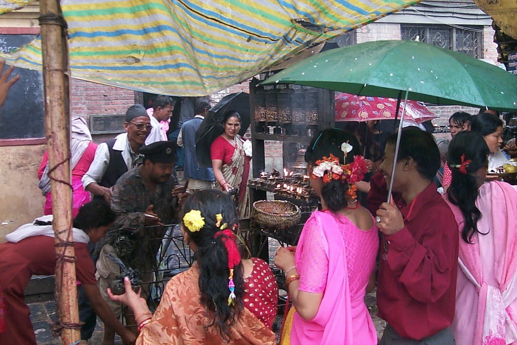 People in colorful traditional attire gather around a ritual flame under tarps and umbrellas, possibly at a religious or cultural event, with a hint of rain.