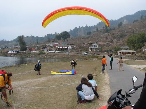 A paraglider prepares to take off from a grassy field near a river and houses, with onlookers watching and another paraglider's gear on the ground.