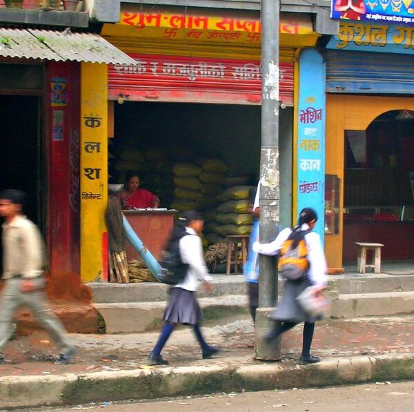Vibrant street scene with people walking past colorful storefronts displaying Nepali script, suggesting a location in Nepal.
