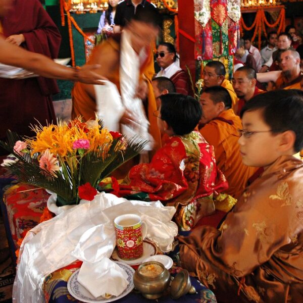 Monks in traditional attire seated in a crowded room with a young boy in ornate clothing, a table with flowers in front, and activity blur from movement.