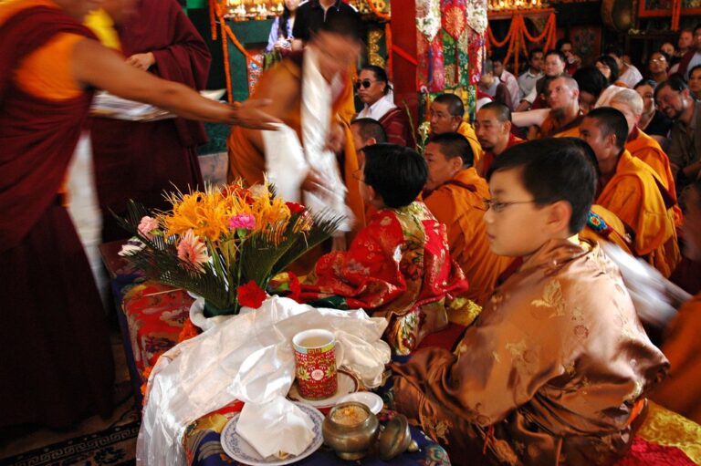 Monks in traditional attire seated in a crowded room with a young boy in ornate clothing, a table with flowers in front, and activity blur from movement.