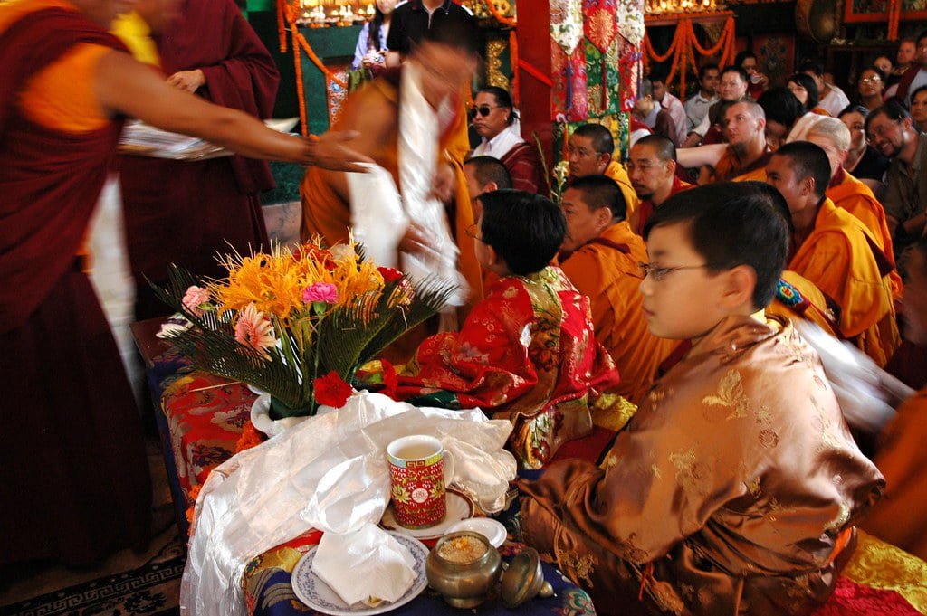Monks in traditional attire seated in a crowded room with a young boy in ornate clothing, a table with flowers in front, and activity blur from movement.