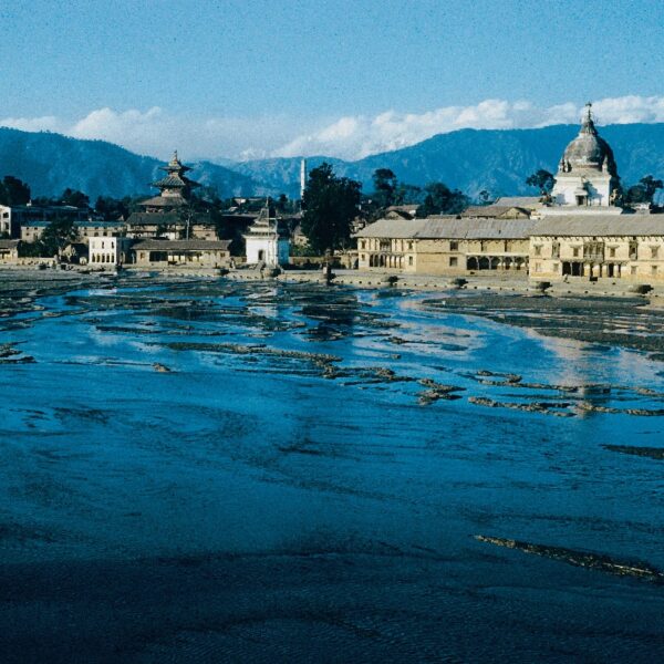 A historic temple complex with multiple tiered roofs beside other traditional buildings, set against a backdrop of distant mountains with an expansive, sunlit, muddy field in the foreground.