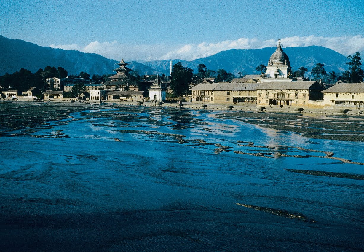A historic temple complex with multiple tiered roofs beside other traditional buildings, set against a backdrop of distant mountains with an expansive, sunlit, muddy field in the foreground.