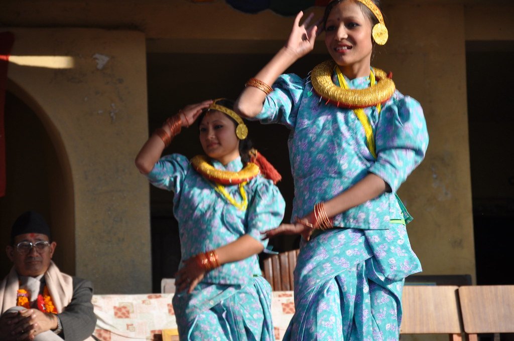 Two women performing a traditional dance in colorful attire with a seated man watching in the background.