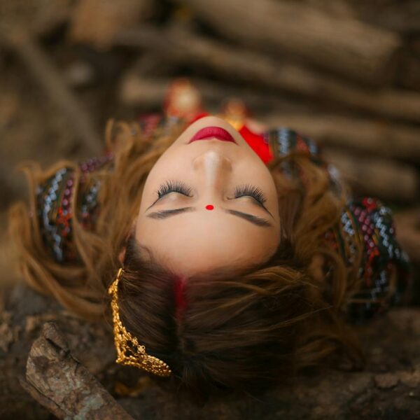 A woman with closed eyes lies on a bed of twigs and leaves, wearing a red bindi, ornate earrings, and colorful embroidered clothing.
