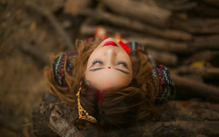 A woman with closed eyes lies on a bed of twigs and leaves, wearing a red bindi, ornate earrings, and colorful embroidered clothing.