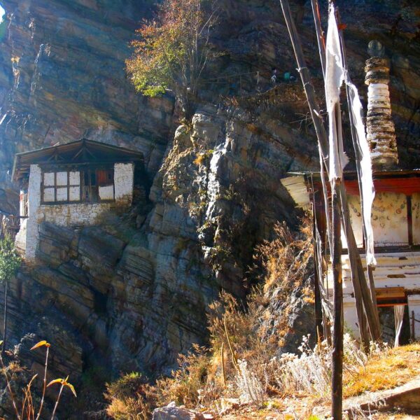 A traditional stone-built house with a wooden roof perched precariously on a steep rocky cliff, flanked by prayer flags and a Buddhist stupa-like structure, in a clear and sunny setting.