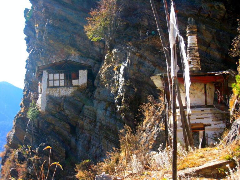 A traditional stone-built house with a wooden roof perched precariously on a steep rocky cliff, flanked by prayer flags and a Buddhist stupa-like structure, in a clear and sunny setting.