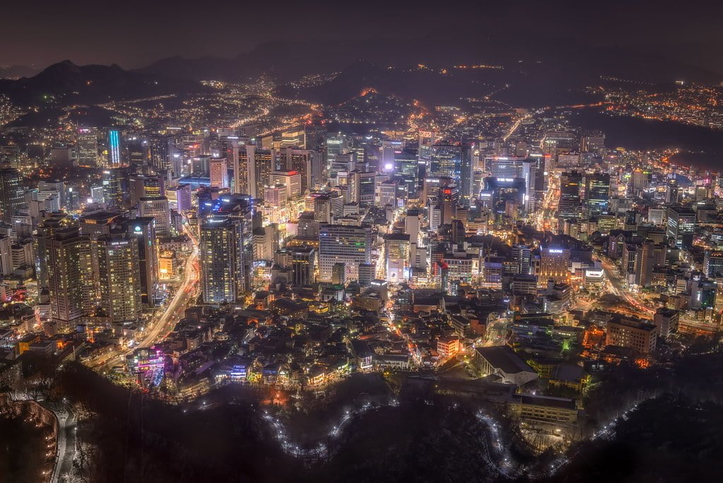 A nocturnal aerial view of a densely populated city with bright lights, towering skyscrapers, and snaking illuminated roadways, surrounded by hilly terrain.
