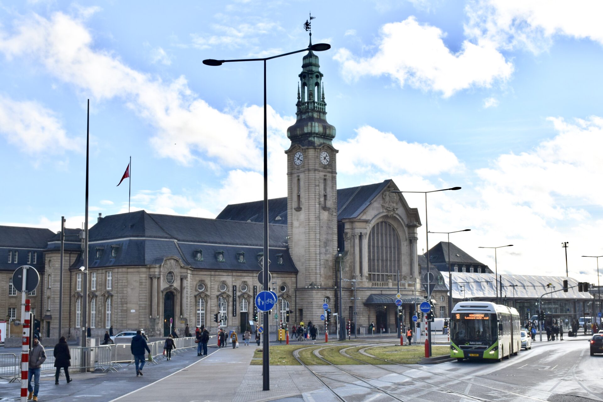 A busy street scene with pedestrians, a modern green tram, and vehicles in front of a historic train station with a prominent clock tower under a partly cloudy sky.
