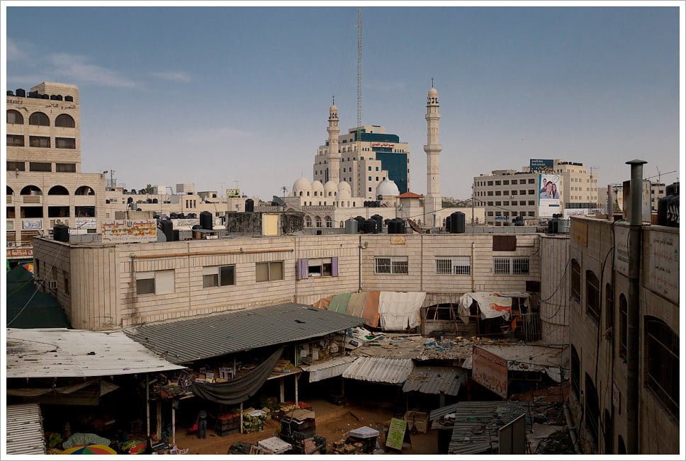 An urban landscape with multi-story buildings, a mosque with a tall minaret, and a crowded market with makeshift stalls.