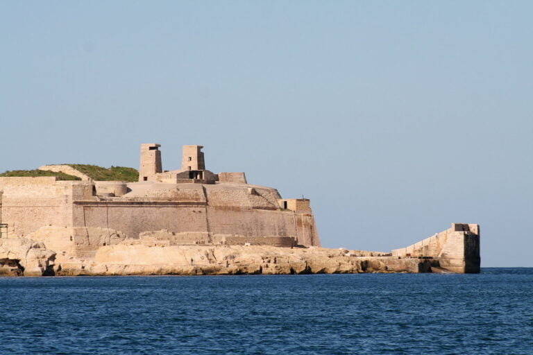 A coastal fortress with intact walls and minimalistic towers stands on a rocky promontory against a clear blue sky, viewed from the sea.