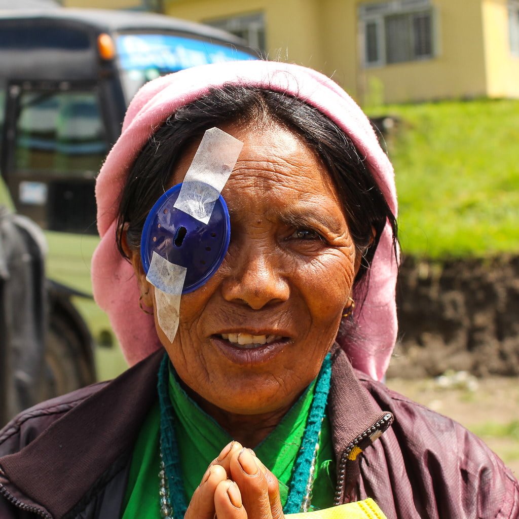 An elderly woman with a patched eye smiling at the camera and wearing traditional attire with a pink headscarf and a green scarf around her neck, with a bus and a building in the blurred background.