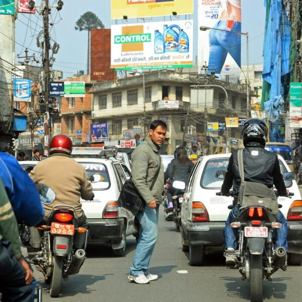 A busy street scene with motorcycles, cars, and a pedestrian crossing the road amidst traffic, with various commercial signboards in the background.