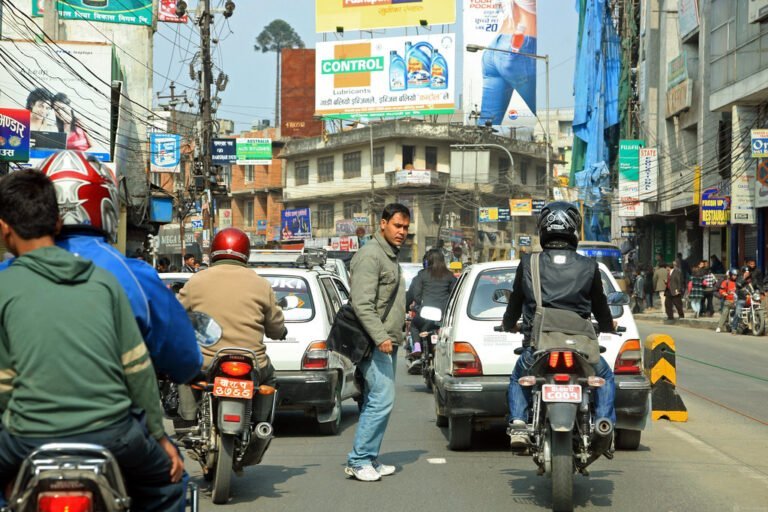 A busy street scene with motorcycles, cars, and a pedestrian crossing the road amidst traffic, with various commercial signboards in the background.