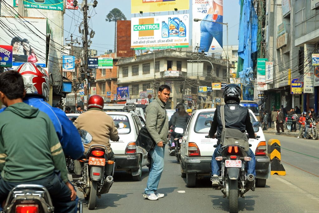 A busy street scene with motorcycles, cars, and a pedestrian crossing the road amidst traffic, with various commercial signboards in the background.