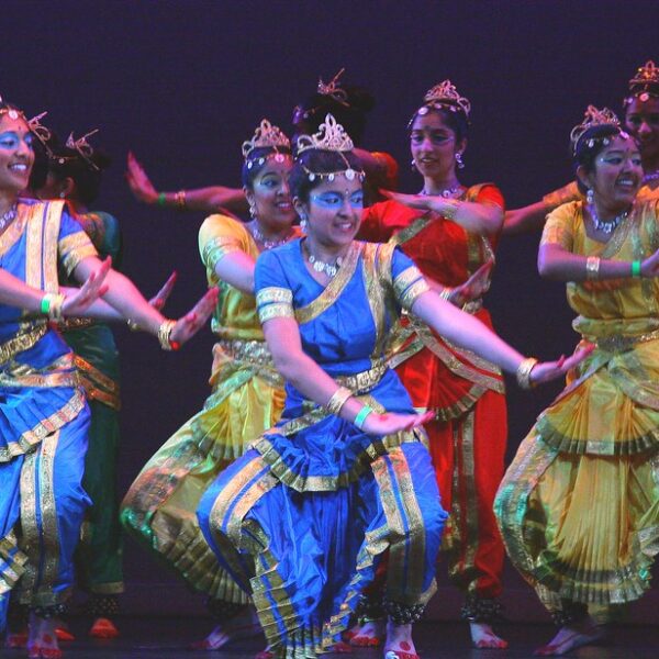 A group of six female Indian classical dancers performing on stage, wearing colorful traditional costumes and headdresses, with dynamic poses and expressions.
