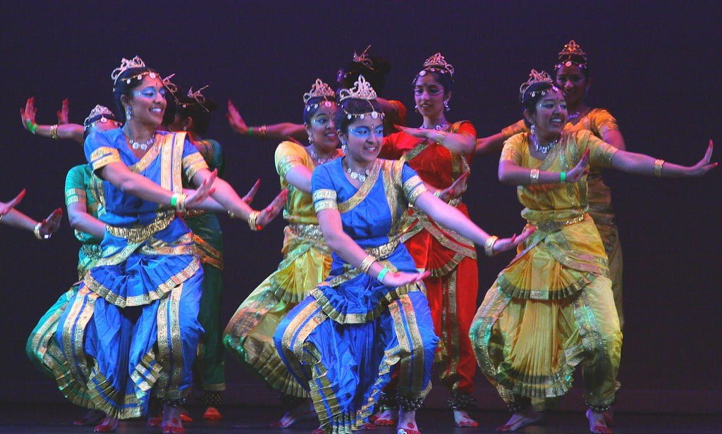 A group of six female Indian classical dancers performing on stage, wearing colorful traditional costumes and headdresses, with dynamic poses and expressions.