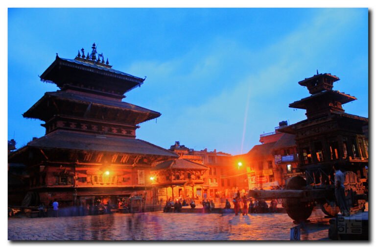 An evening view of two traditional multi-tiered temples at Durbar Square with people gathered and walking around in the blue hour light.