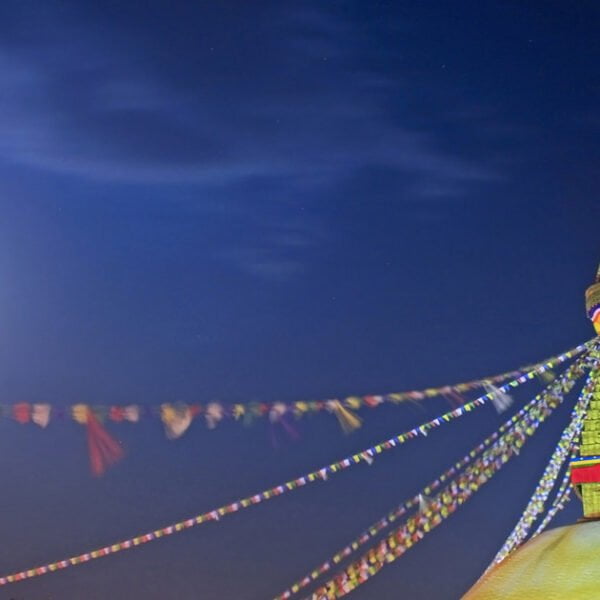 A brightly illuminated stupa at night with prayer flags radiating from the top against a dark blue sky, with a full moon slightly visible on the left.