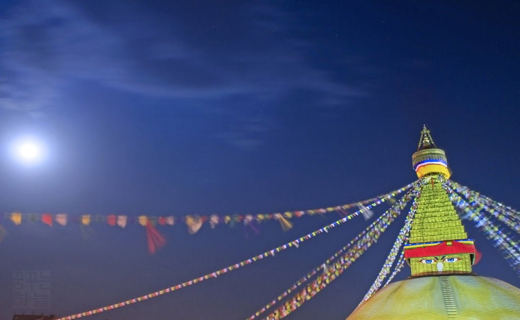 A brightly illuminated stupa at night with prayer flags radiating from the top against a dark blue sky, with a full moon slightly visible on the left.