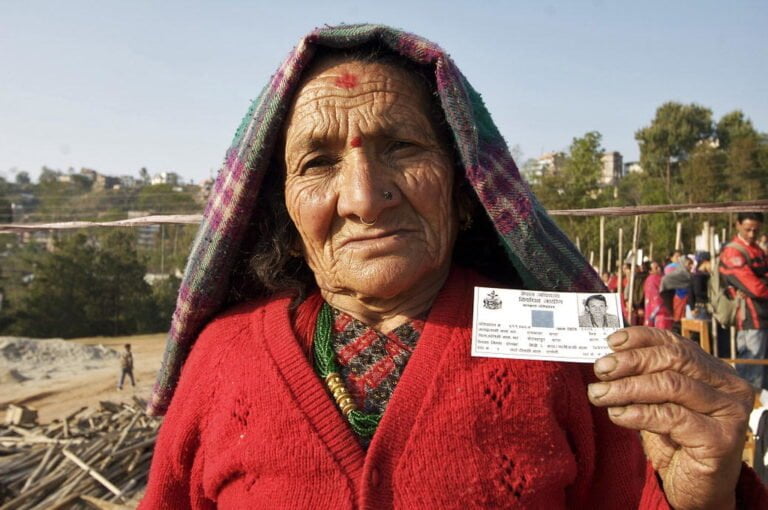 An elderly woman with a red sweater and a green patterned shawl over her head, holding an identification card outdoors, with construction material and other people in the background.