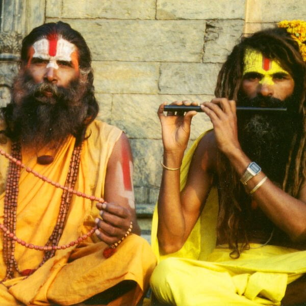 Two men in traditional saffron robes with painted foreheads sitting in front of an old stone structure; one holds prayer beads while the other plays a flute.