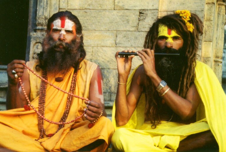 Two men in traditional saffron robes with painted foreheads sitting in front of an old stone structure; one holds prayer beads while the other plays a flute.