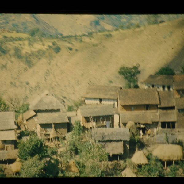 A rustic village with thatched and tiled roof houses nestled in the hills, captured in an old, somewhat blurry photograph.