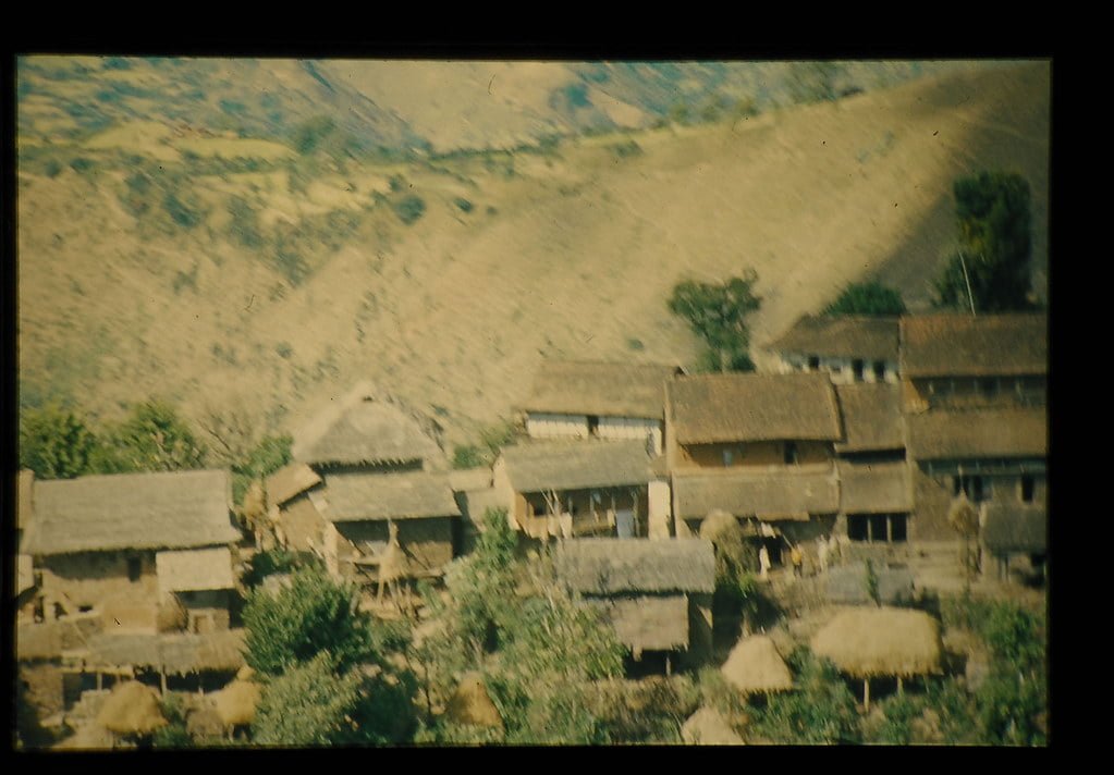 A rustic village with thatched and tiled roof houses nestled in the hills, captured in an old, somewhat blurry photograph.