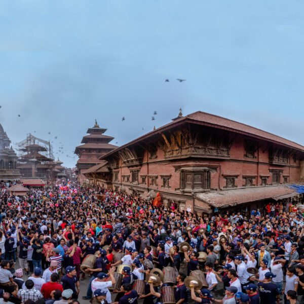 A panoramic view of a bustling festival at a historical square, with a dense crowd of people gathered around a towering chariot and traditional buildings, under a twilight sky.
