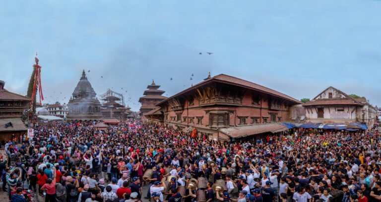 A panoramic view of a bustling festival at a historical square, with a dense crowd of people gathered around a towering chariot and traditional buildings, under a twilight sky.