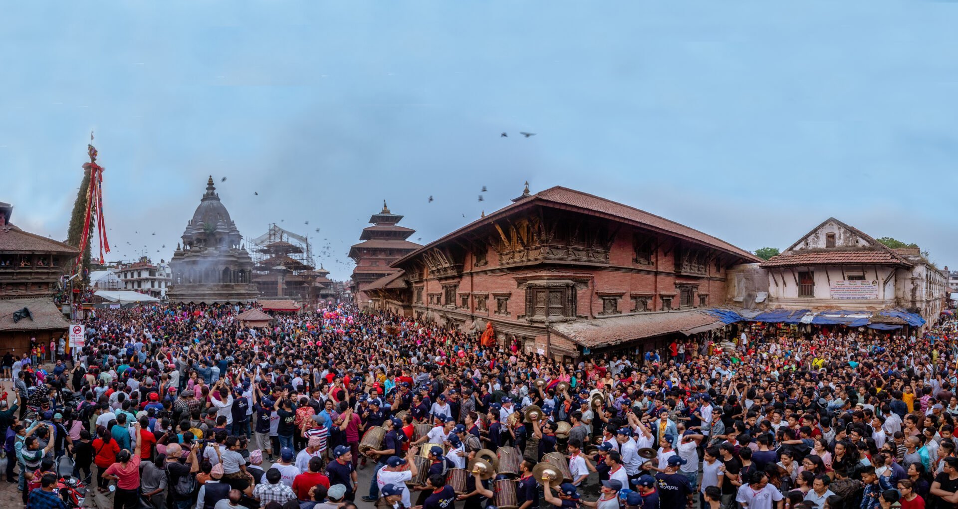 A panoramic view of a bustling festival at a historical square, with a dense crowd of people gathered around a towering chariot and traditional buildings, under a twilight sky.