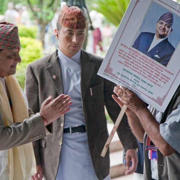 Two men, one holding a placard with a photo and text tribute, possibly at a commemorative event, with people in the background.