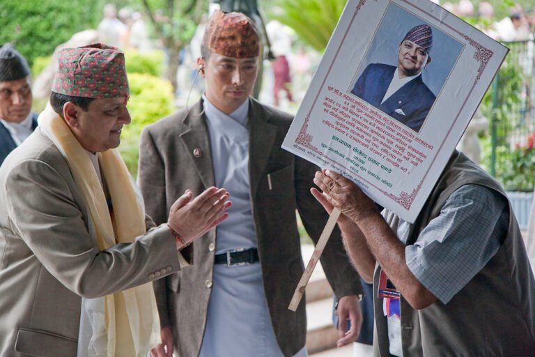 Two men, one holding a placard with a photo and text tribute, possibly at a commemorative event, with people in the background.