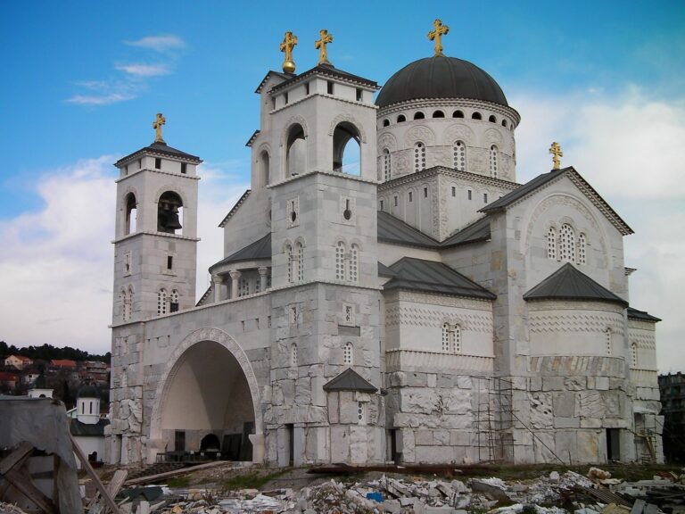 A large, ornate Orthodox church with a black dome and gold crosses under construction, surrounded by building materials, with a blue sky in the background.